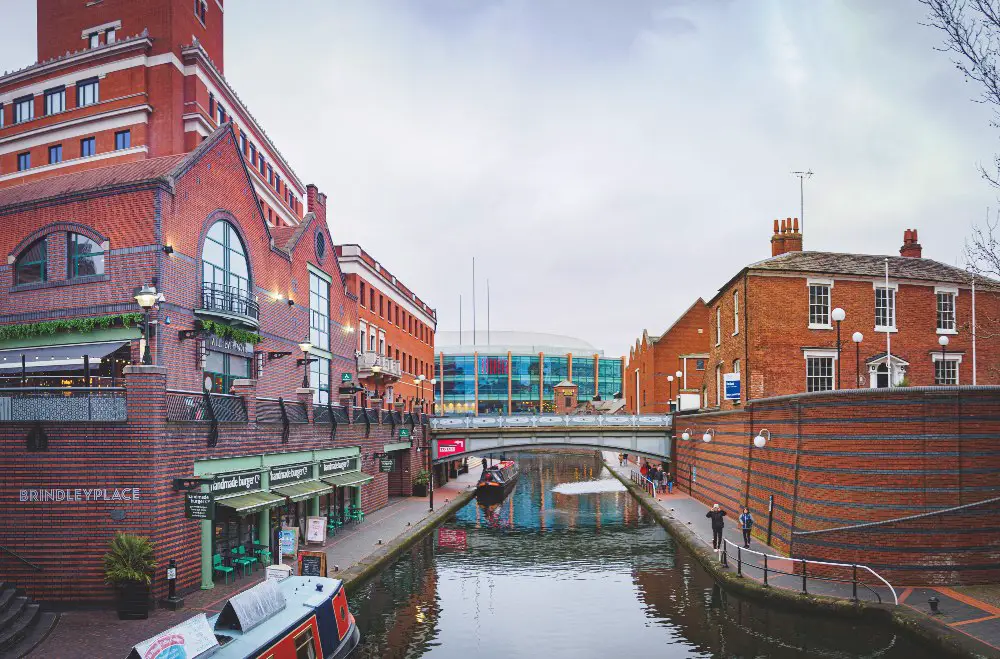 A scenic view of Brindleyplace in Birmingham, UK, featuring a canal running between two brick buildings. On the left, there is a large red-brick building with the sign 'Brindleyplace' visible, and a restaurant with outdoor seating by the canal. On the right, there is a smaller, traditional brick building. A narrowboat is moored along the canal, and a pedestrian bridge crosses the canal in the background. The atmosphere is calm, with a few people walking along the canal path.
