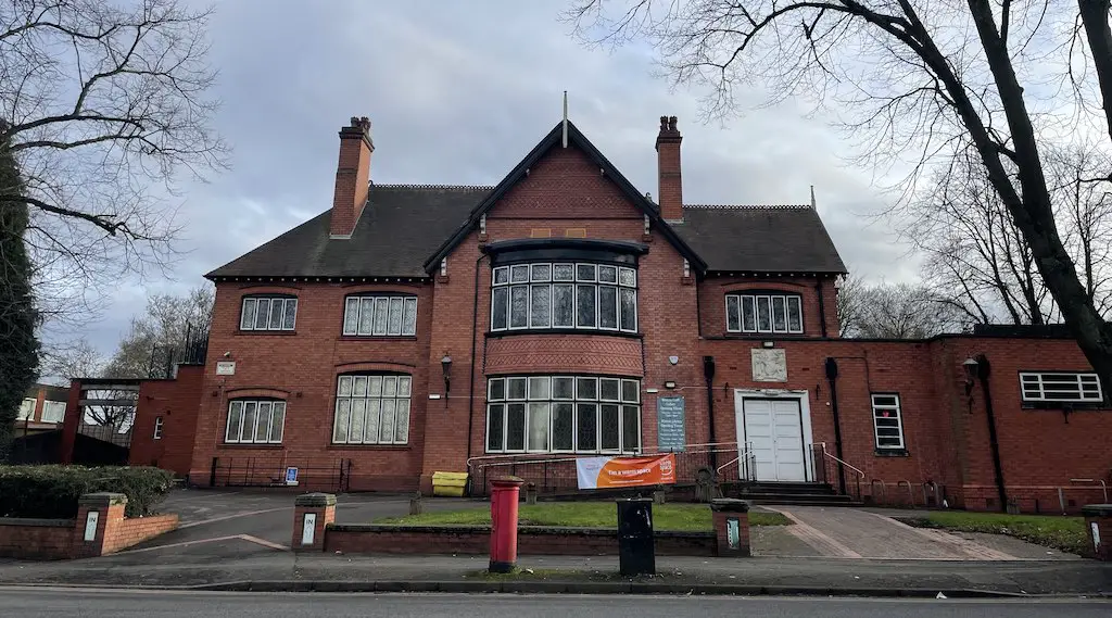 Bilston Craft Gallery, a local art gallery showcasing the work of talented artists, is housed in a historic red brick building with a gabled roof, tall chimneys, black and white framed windows and doors, and a prominent bay window. The building is surrounded by trees and has a paved driveway leading to the entrance.