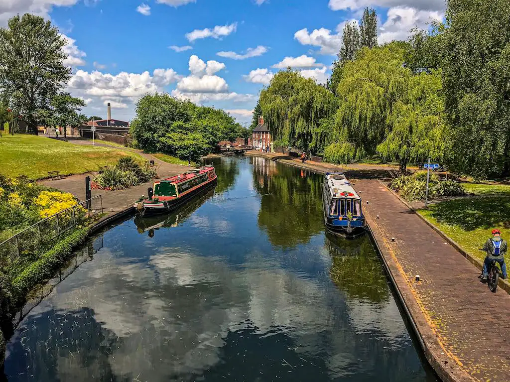 A peaceful canal scene on a sunny day with vibrant blue skies and fluffy white clouds reflected in the still water. Narrowboats are docked along the sides of the canal, bordered by lush greenery, large weeping willow trees, and a cobblestone path. A cyclist in the distance enjoys the tranquil environment.