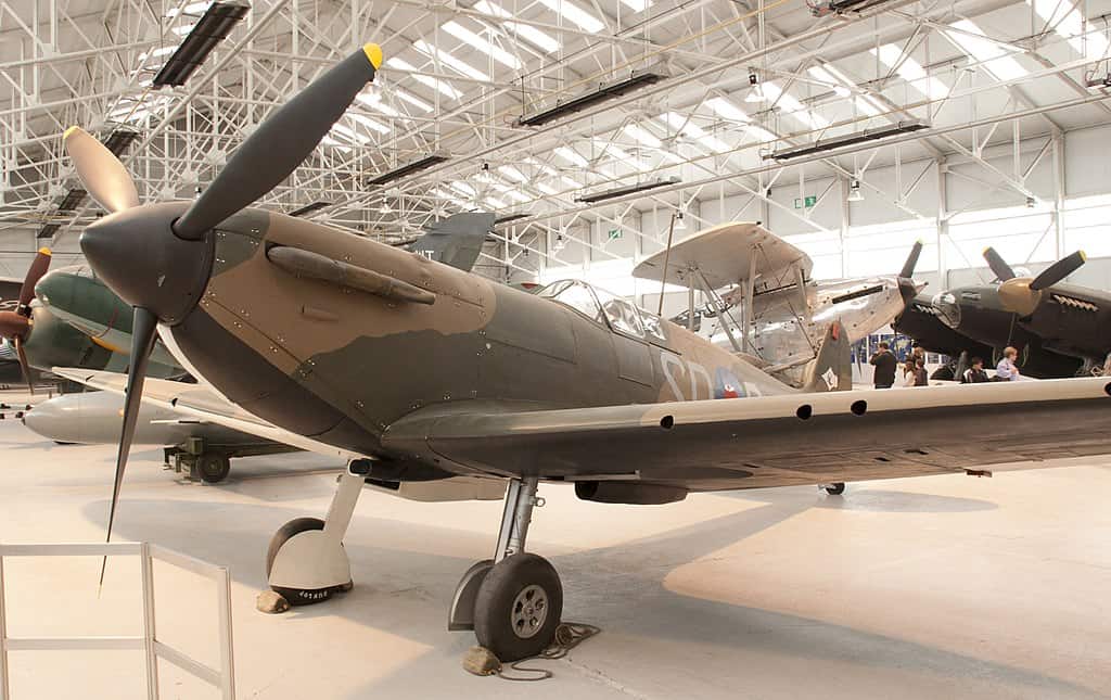 The image shows a Spitfire aircraft displayed in a museum. The plane has a sleek, streamlined body with a propeller at the front and camouflage paint in shades of brown and green. It is positioned inside a large, well-lit hangar with other vintage aircraft visible in the background. The Spitfire's distinctive elliptical wings and landing gear are clearly visible, highlighting its iconic World War II design.