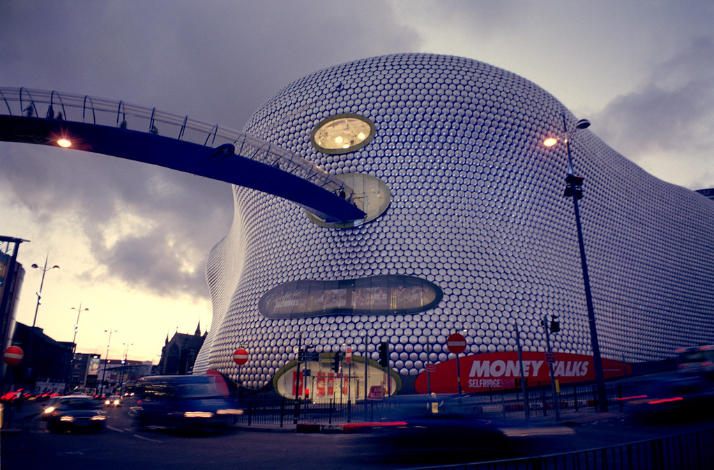 large silver-scaled fluid modern building with round large windows and a long blue tunnel bridge extending out over a road below and off the left side of the picture.