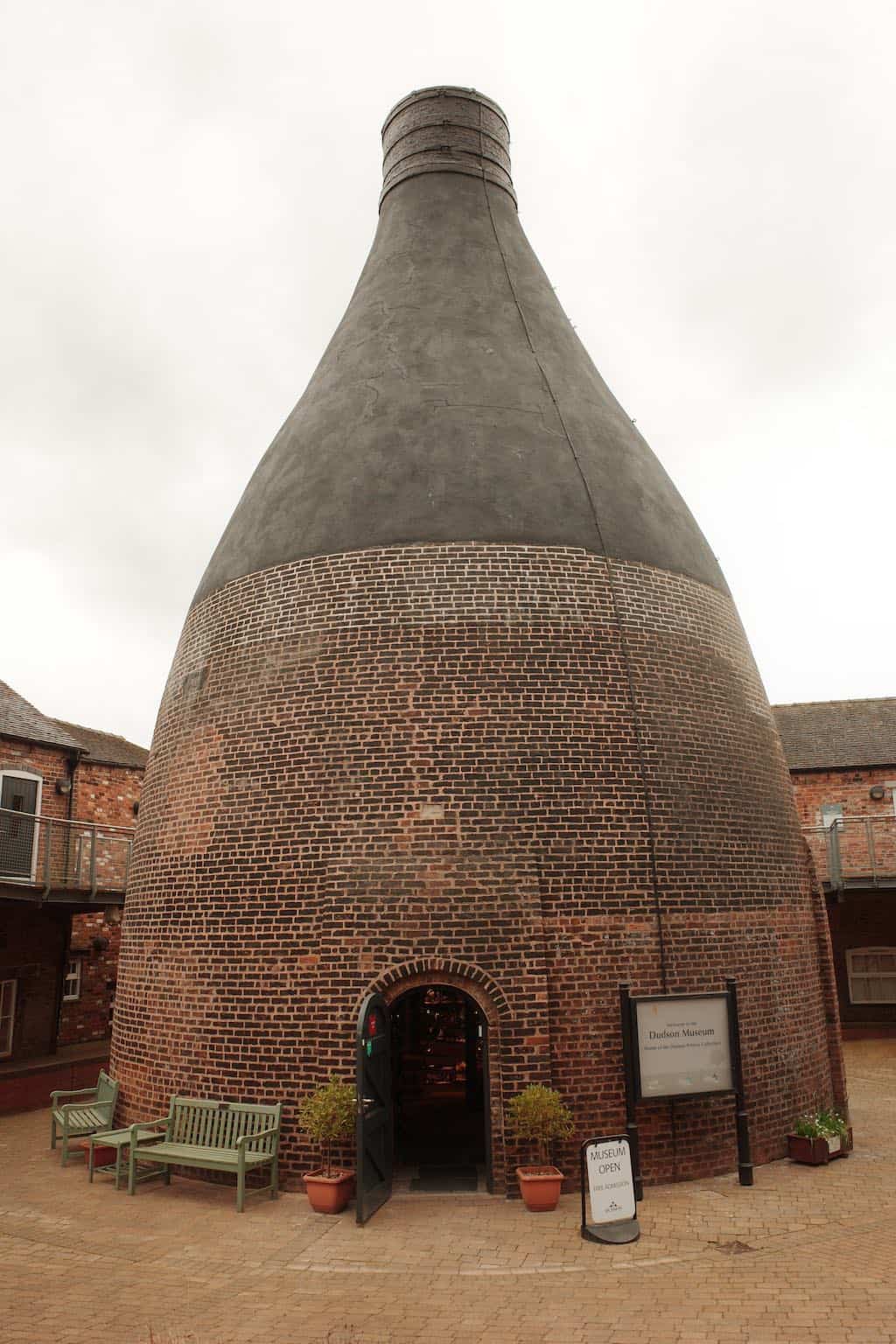 large brick bottle kiln with an open door at the base. surrounded by factory buildings