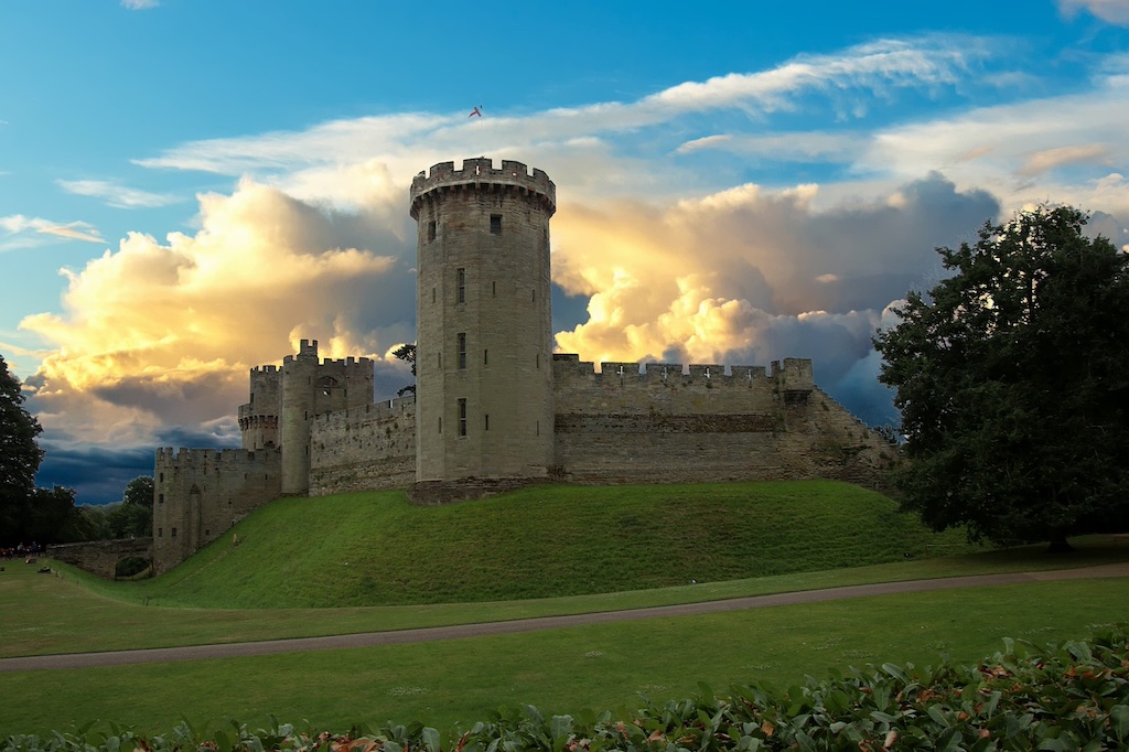 large medieval castle on a grassy hill with clouds in the background and trees at the side