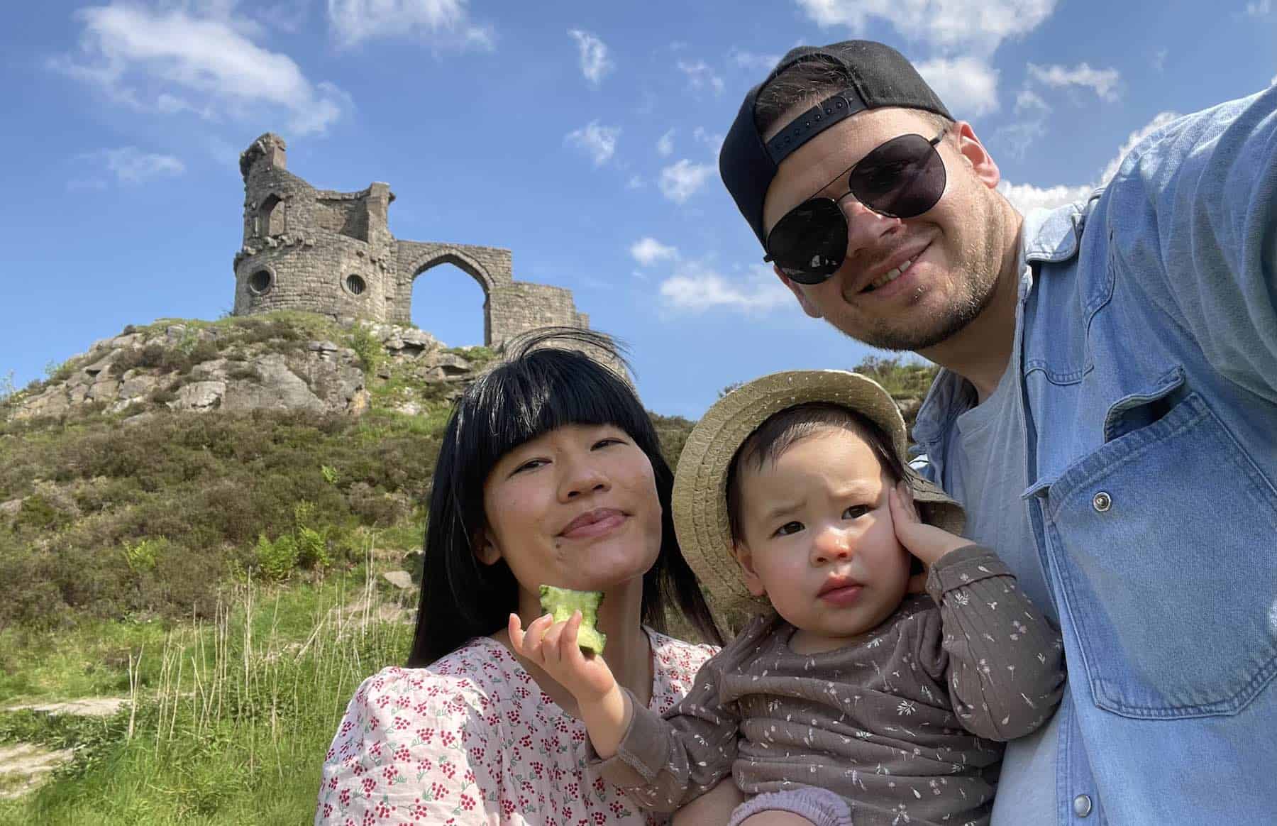 smiling young couple with a toddler, posing in front of a castle ruin on a hill.