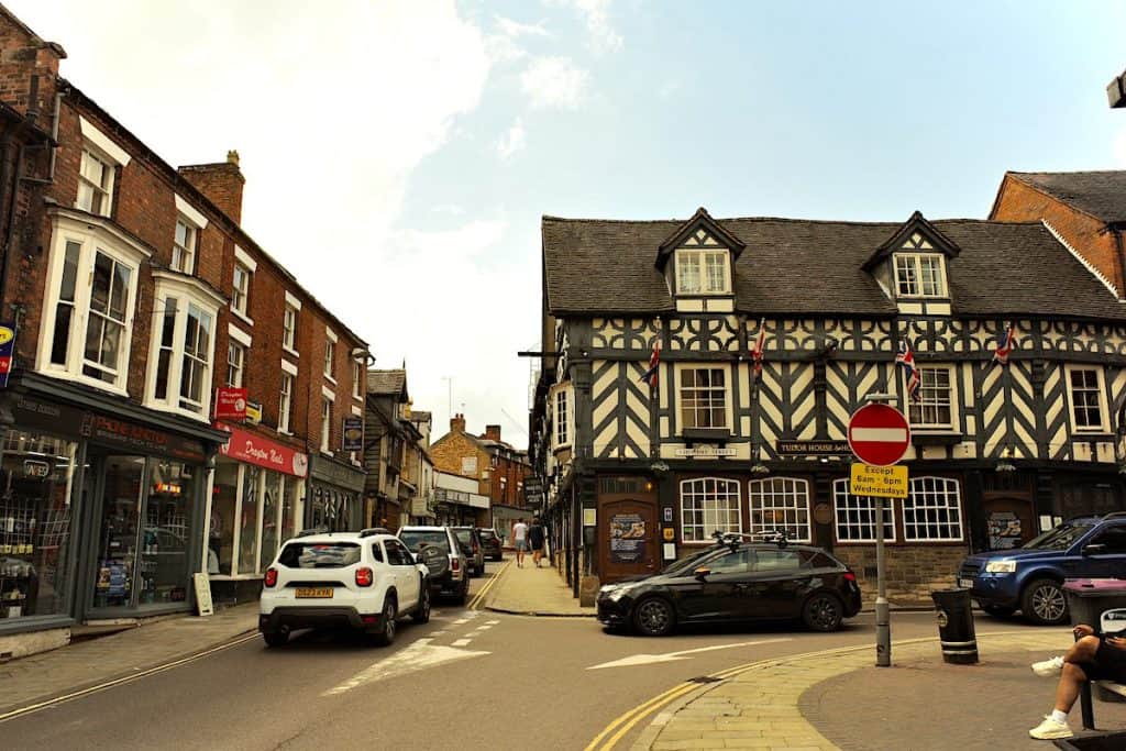A charming street scene in a historic town. On the right is a large half-timbered building with Union Jack flags, likely a pub. The street is lined with a mix of brick and Tudor-style buildings. Cars are parked along the street, and a few pedestrians can be seen walking on the sidewalk.