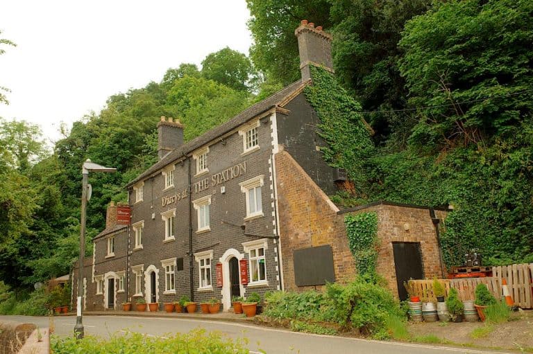 Historic brick building with "D'arcys at The Station" signage on the facade. A former railway station converted into a restaurant, nestled against a wooded hillside with ivy growing up its walls and terracotta planters lining the entrance.