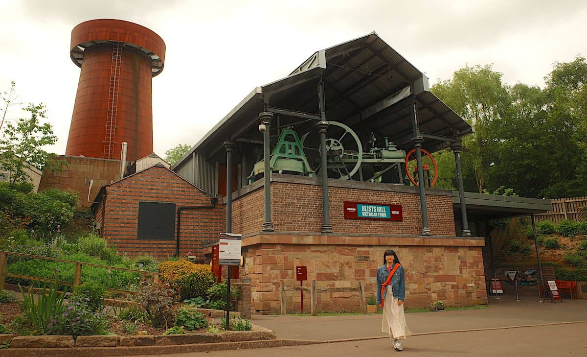 a young lady standing at the front entrance of blists hill victorian town, with large chimney in background and giant 19th century factory machines on display