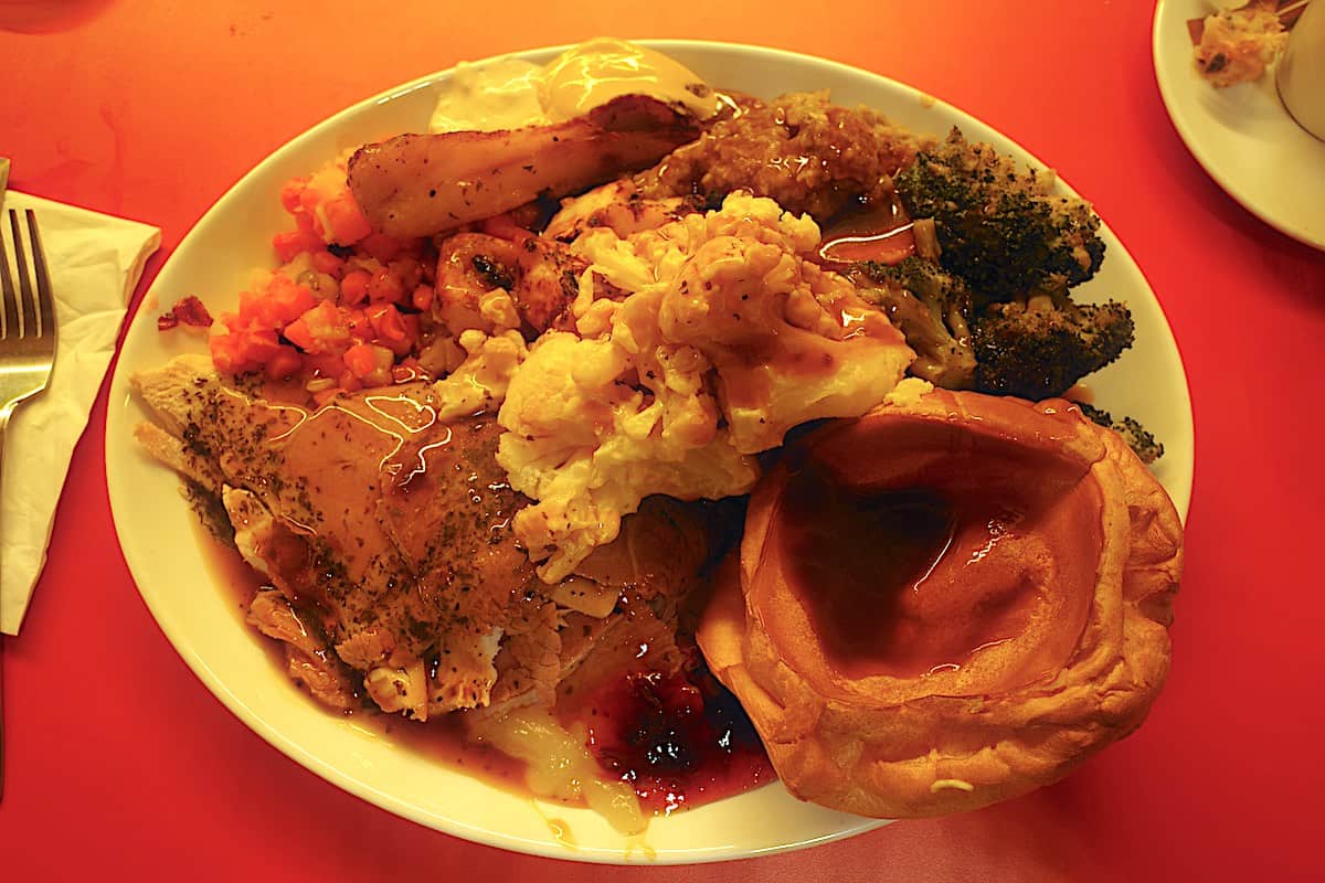 large white plate loaded with slices of turkey, ham and beef, along with a yorkshire pudding, vegetables, garnishes of apple, cranberry sauces, horseradish and mustard, with mint gravy on top.