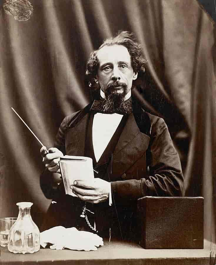 black and white photographic portrait of 19th century novelist charles dickens in a black suit, holding a pen and paper and in thought.
