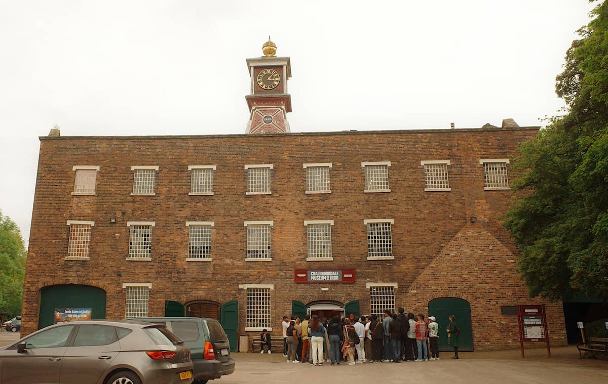 a large victorian, brick factory building with a clock tower on top, many victorian windows on the front and alrge green doors, with people gathering around the entrance, and some parked cars in front.