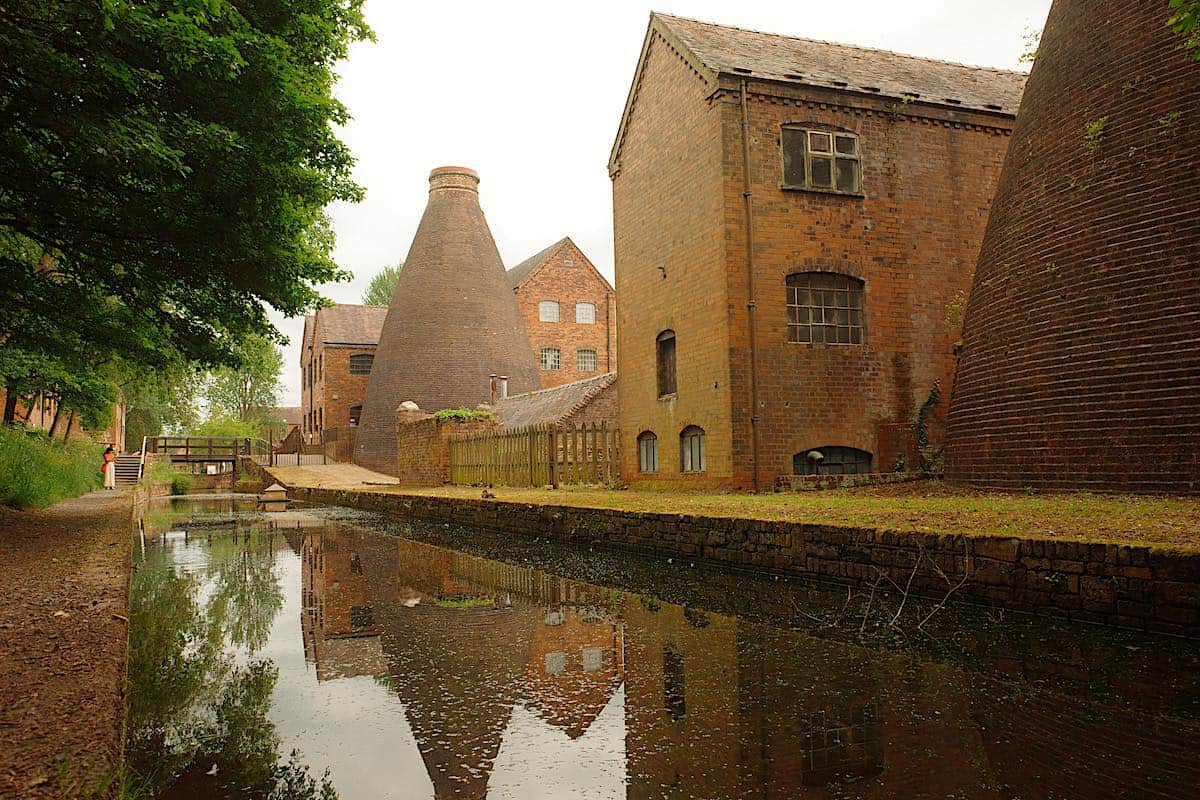 A serene canal lined with trees on one side and red brick industrial buildings on the other, with a distinctive bottle kiln structure reflecting in the water.