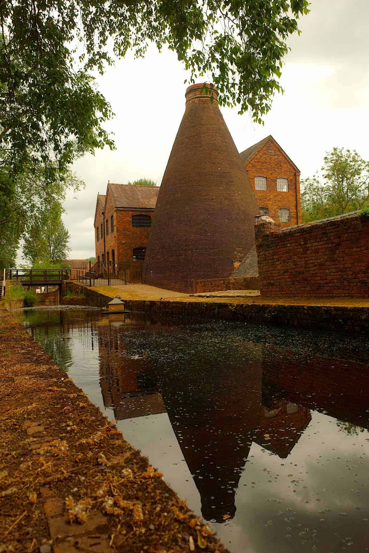view of a large brick bottle kiln with victorian factory buildings around it, and in front, a canal reflecting the buildings, on the other side is a walkway and some trees.