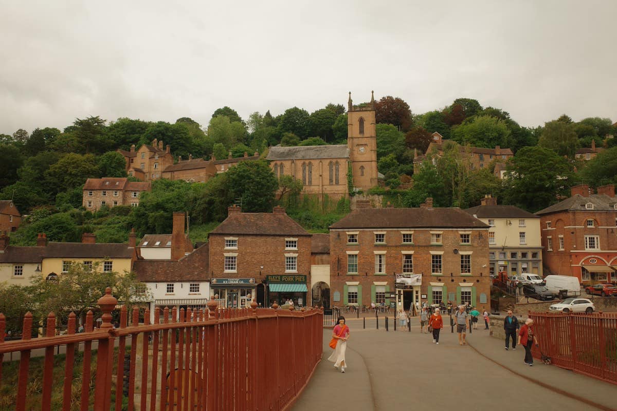 View of a small historic town built on a hillside, seen from a bridge with red railings. A church with a tall tower stands prominently on the hill, surrounded by Georgian and Tudor-style buildings. The town features various shops and businesses at street level, with residential buildings rising up the slope. People are walking across what appears to be a pedestrian bridge in the foreground. The overall scene captures a traditional English market town with buildings from different historical periods.