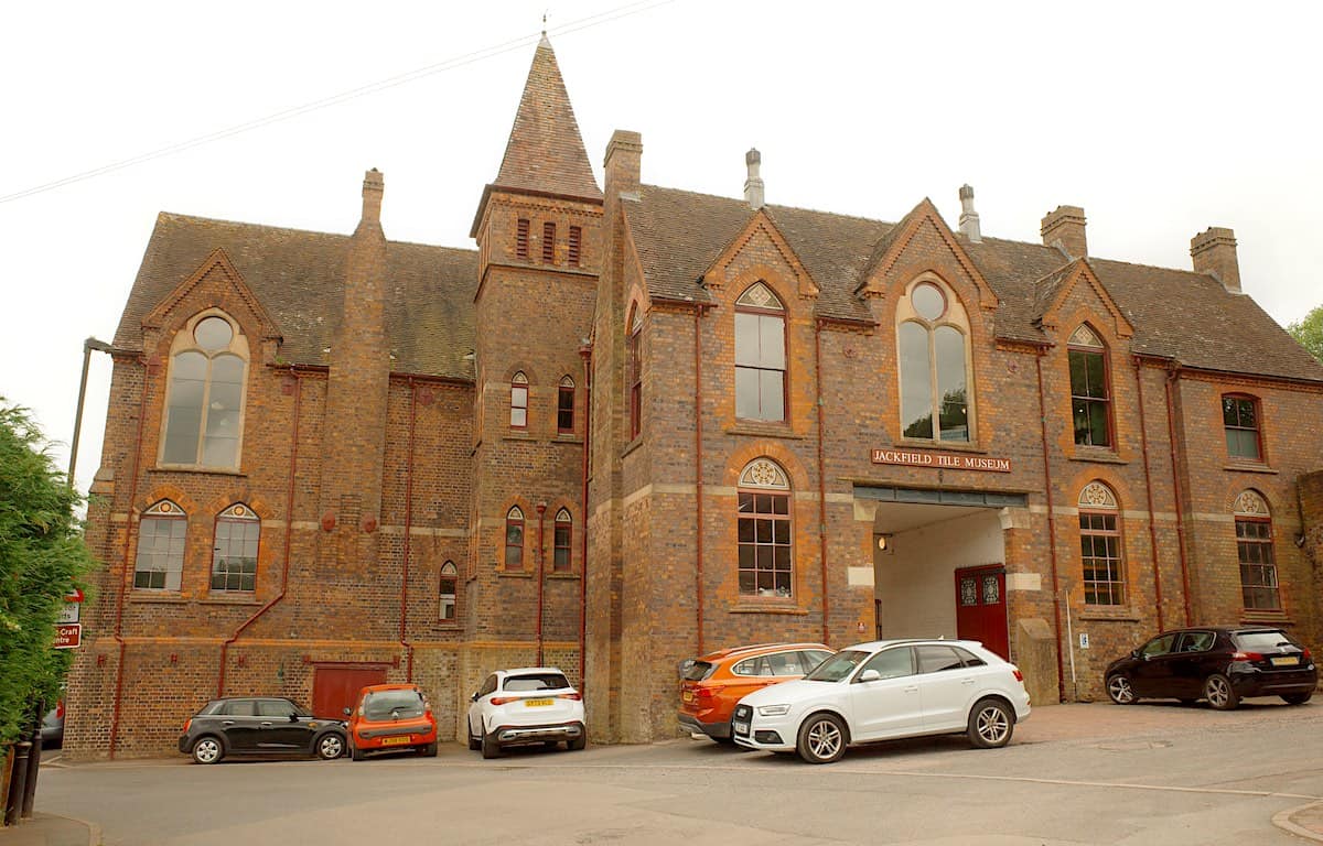 front view of the red brick walled jackfield tile museum, with intricate tiling detail and old, tall victorian windows, with a spire and chimneys lining the roof and museum sign above the entrance, and some cars parked in front of the building