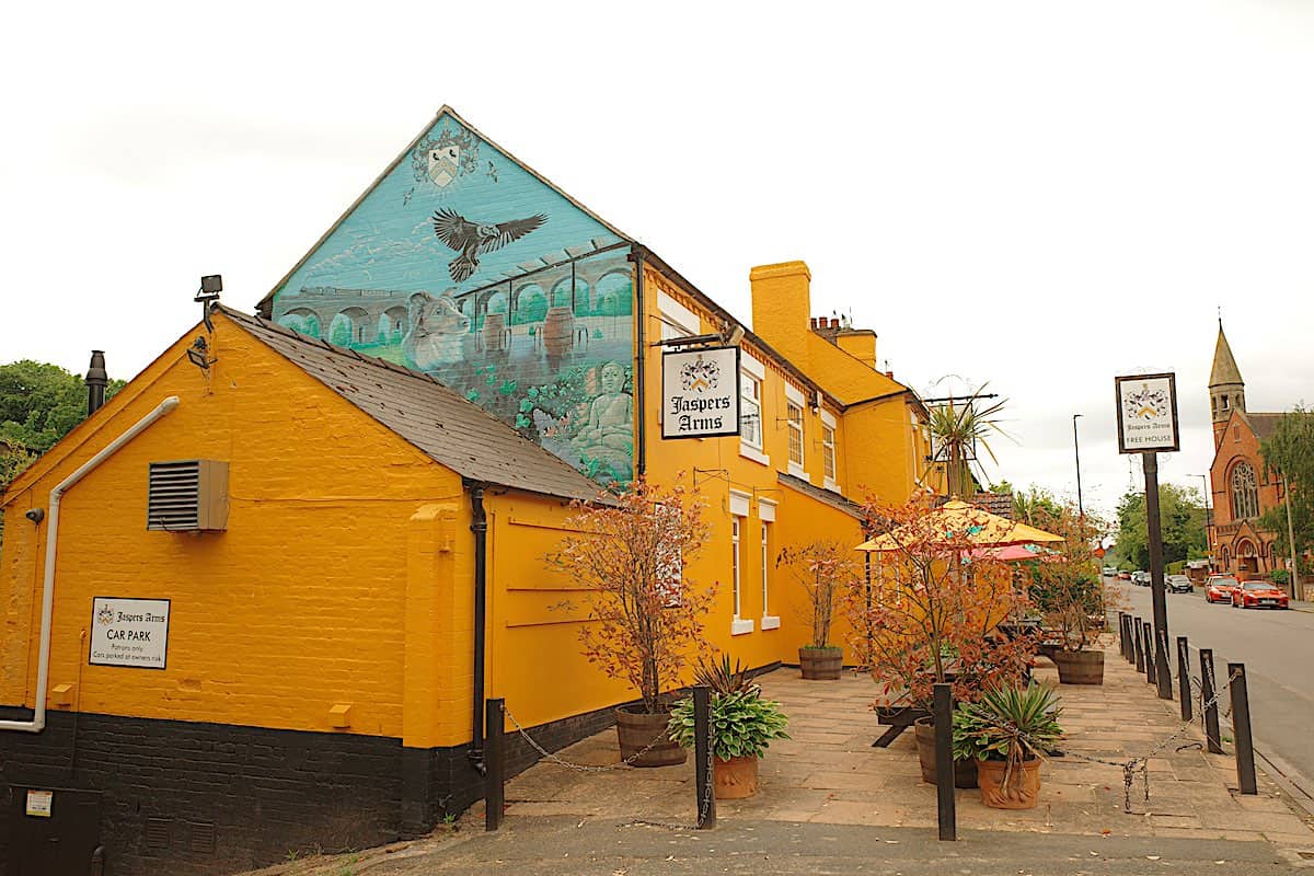 bright yellow painted old building housing the jaspers arms pub, with a church in the distance and trees in the background