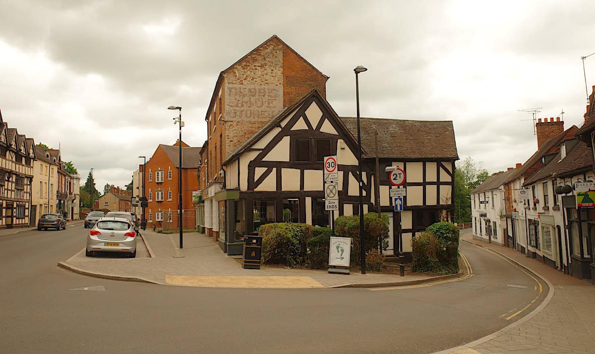 A street corner in a historic town showing half-timbered buildings, brick structures, and a mix of architectural styles. A sign for "Globe Shoes Store" is visible on one building. Cars are parked along the street, which has both pedestrian and vehicle areas.