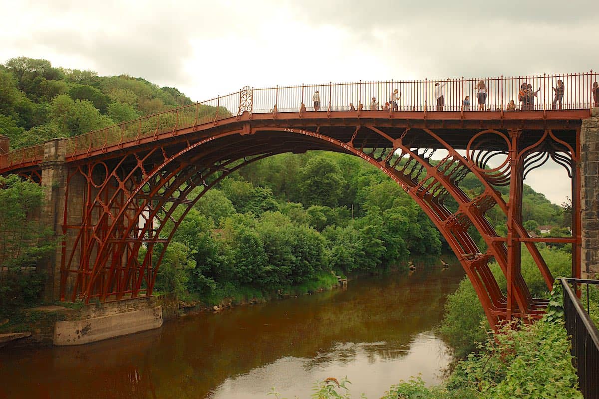 large 18th century, red-painted, cast-iron bridge over a river on a valley with trees on either side and people walking across the bridge