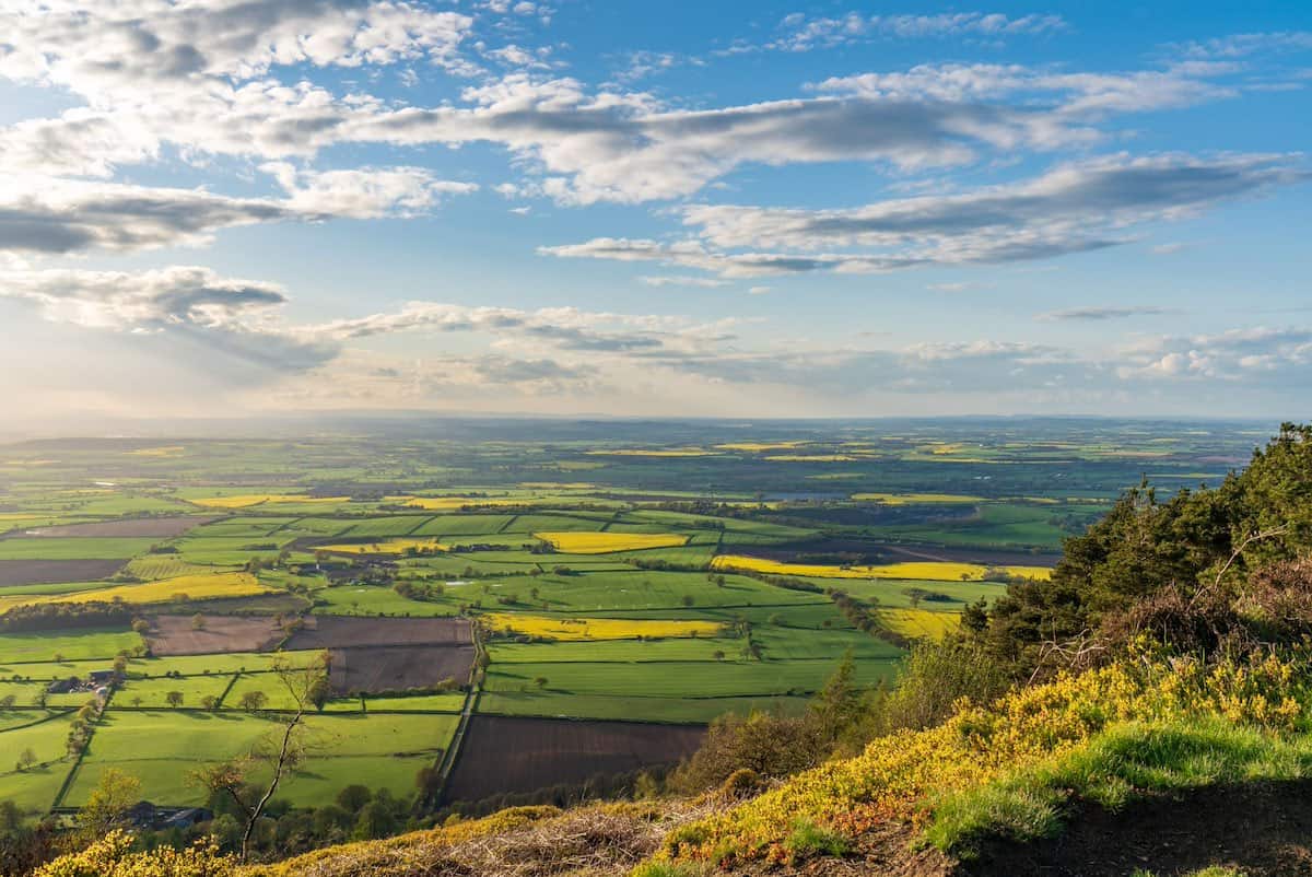 view from the slopes of the wrekin peak, looking down at the green and yellow hues of countryside, and clouds in the sky