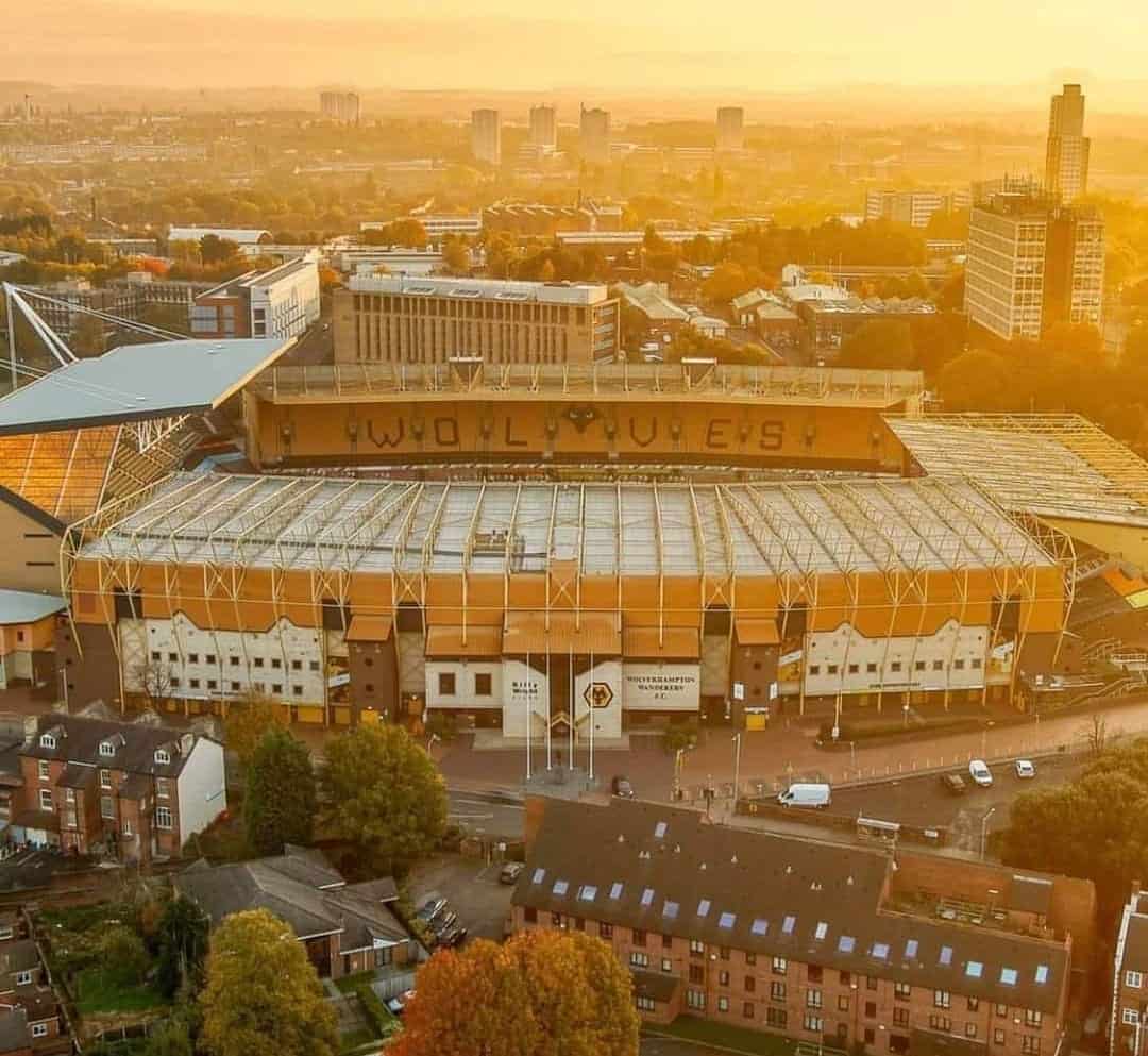 Molineux Stadium, the home of the Wolverhampton Wanderers football club, stands majestically against the backdrop of a sprawling cityscape bathed in the golden hues of a setting sun. The stadium's distinctive architecture and vibrant colors are showcased in this aerial view, capturing the essence of a beloved sporting venue.