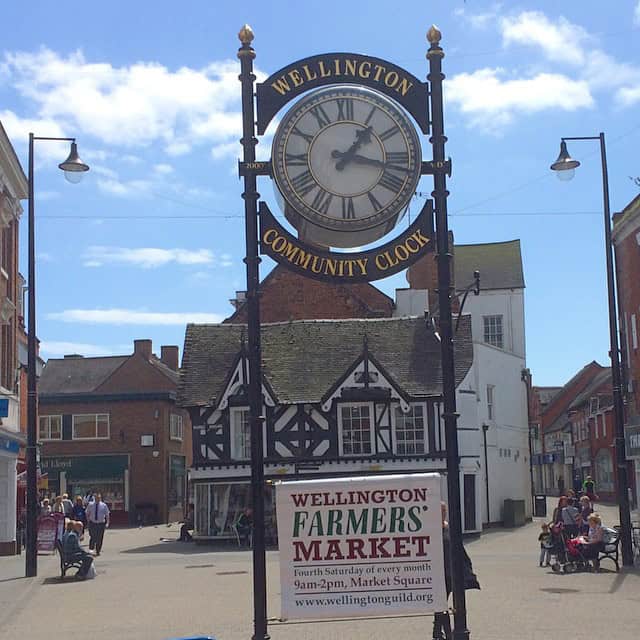 a large clock on poles with the sign around it saying wellington community clock, and a lower sign saying wellington farmers market, and behind it a tudor building in the centre of town, with the high street splitting off around it, with shops along both sides