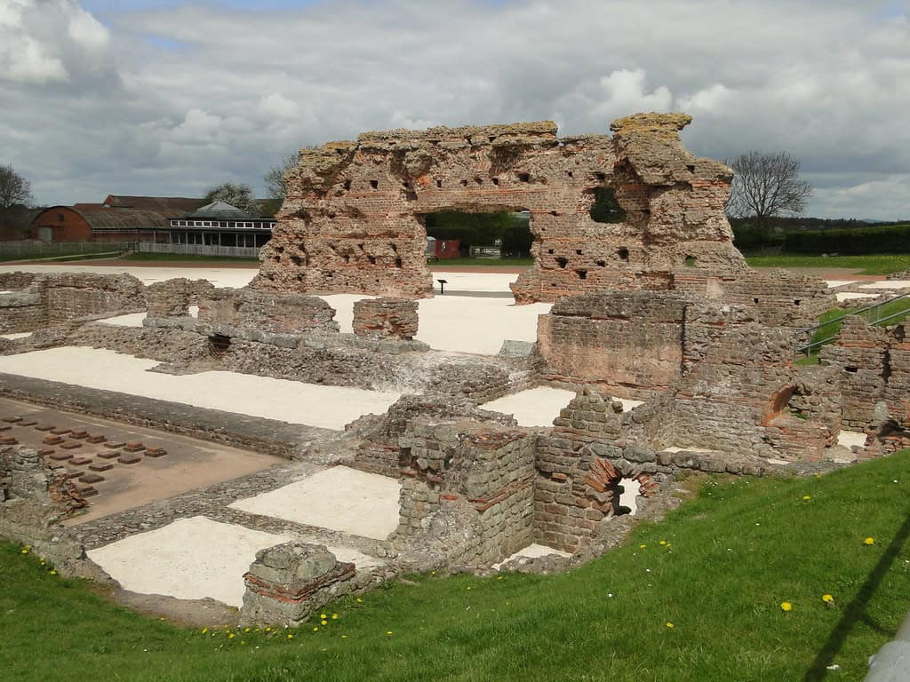 Ruins of an ancient Roman structure, likely a bath complex, with crumbling brick and stone walls. The site features exposed foundations, arched openings, and various rooms or chambers. In the foreground, there's a grassy area with small yellow flowers. The background shows a cloudy sky and some modern buildings, indicating this is an archaeological site preserved within a contemporary setting.