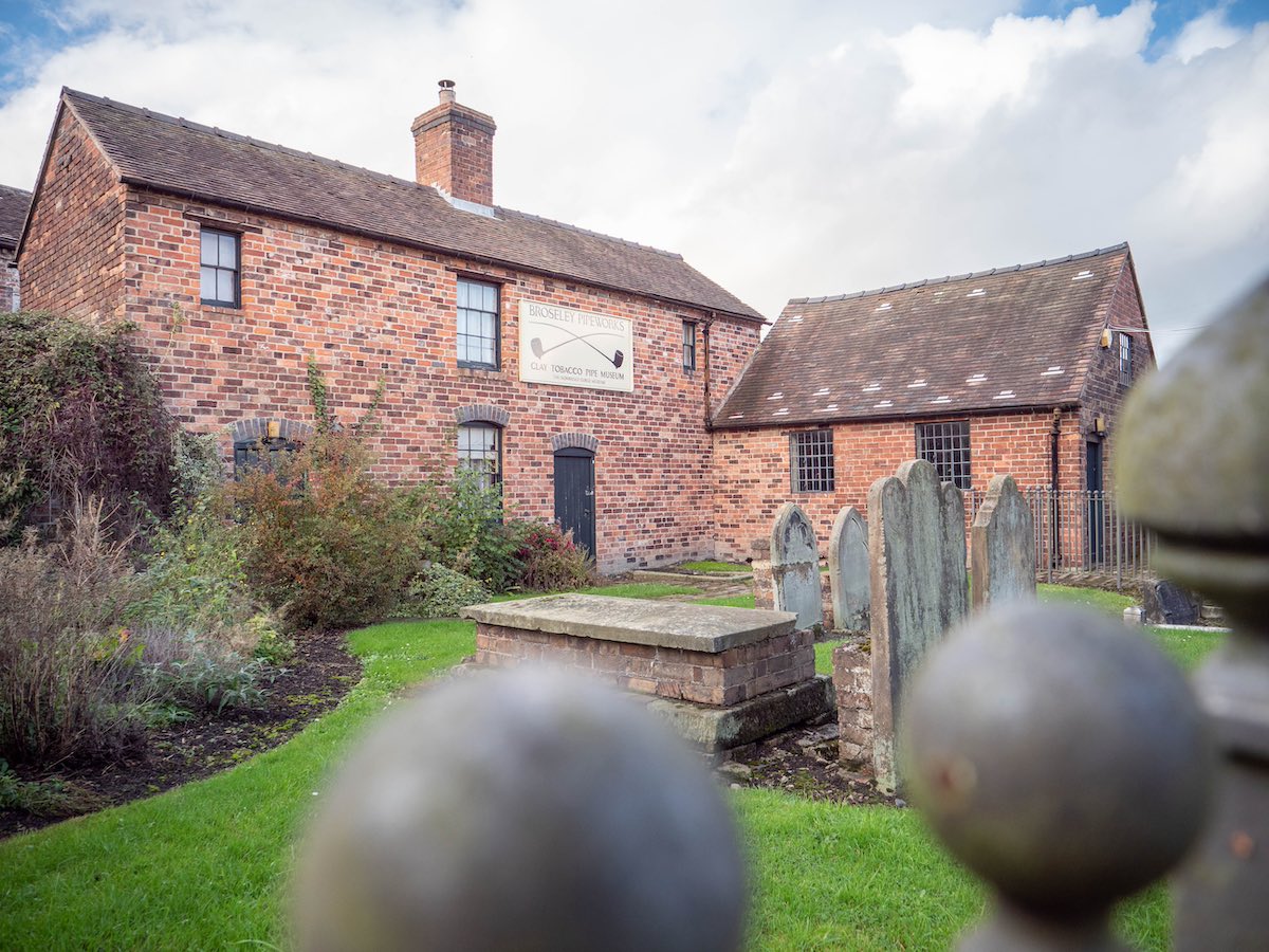 A quaint red brick cottage with a sloping roof, surrounded by a garden and a wooden fence, viewed through a pair of stone gateposts.