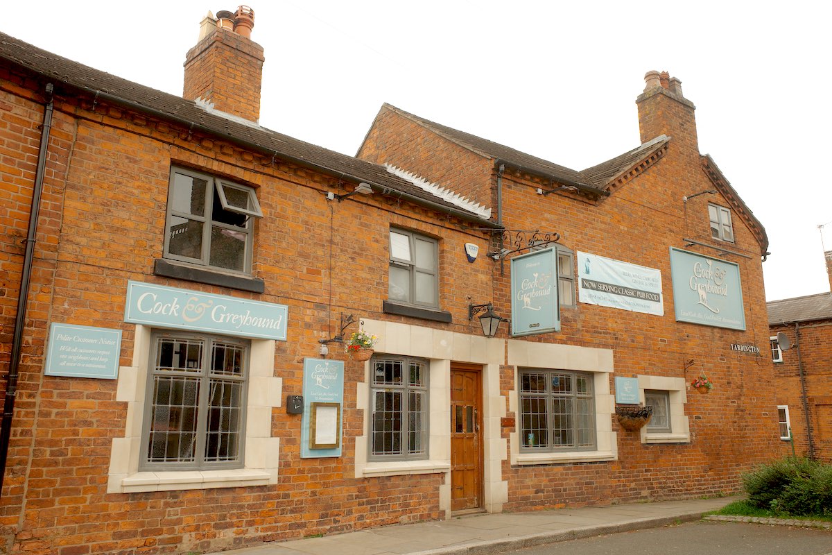 large brick building with pastel blue signage saying the cock and greyhound, and cream-coloured brick framed windows and doors
