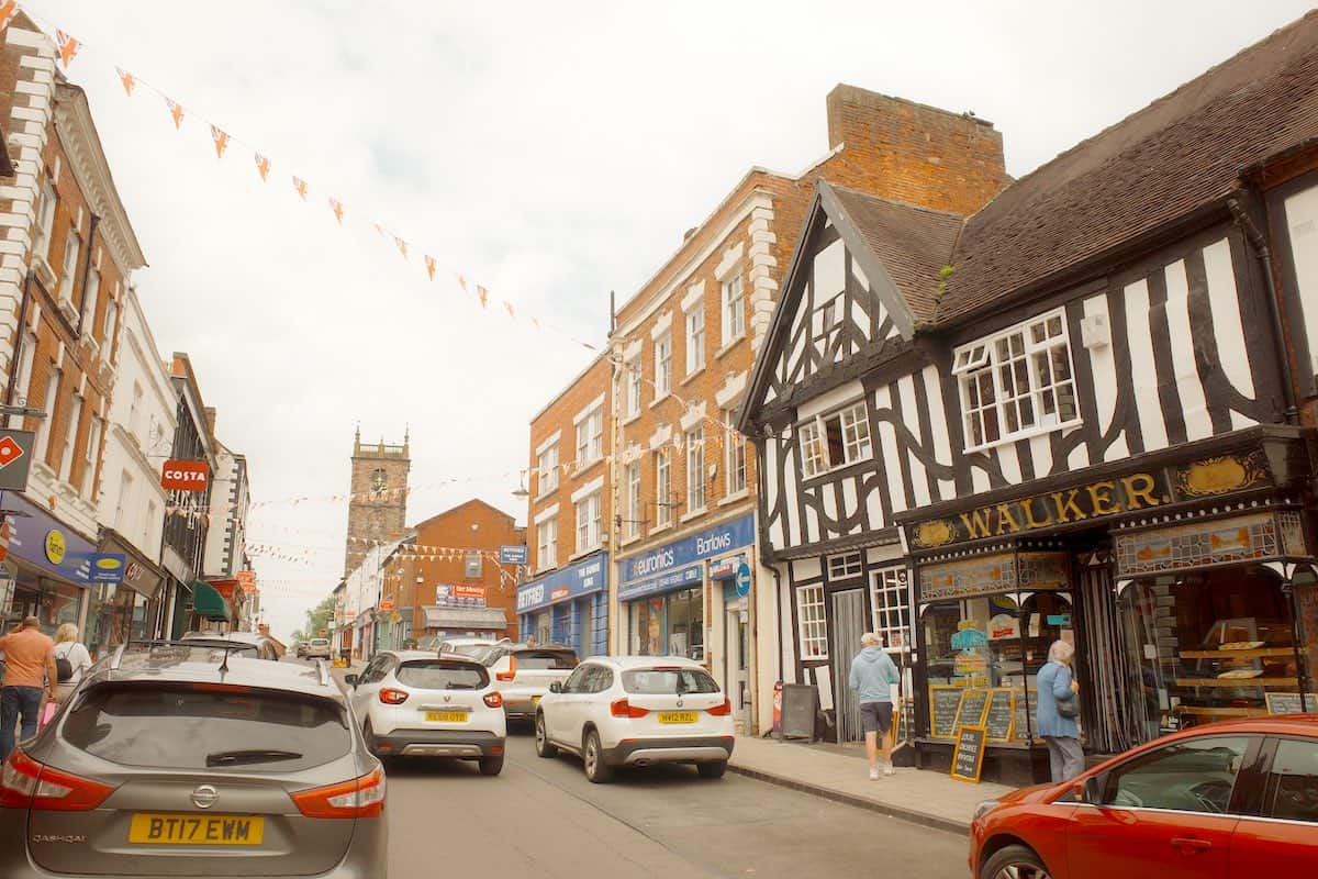 high street with flag bunting criss crossing the street and various medieval, tudor and georgian buildings lining both sides and cars driving through the street