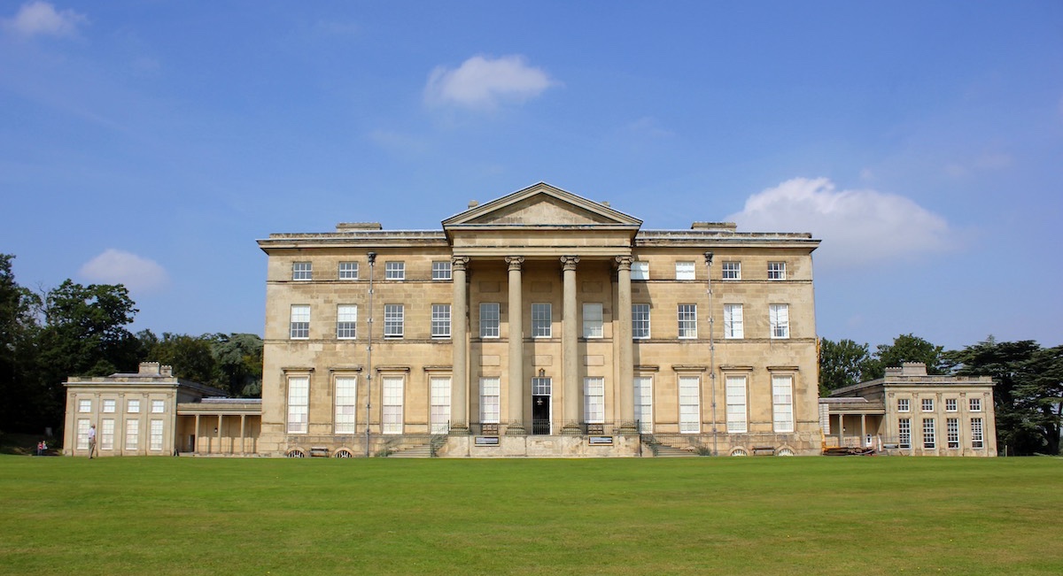The image shows the Mansion at Attingham Park, a grand neoclassical building located in Shropshire, England. The structure features a symmetrical façade with tall columns supporting a triangular pediment above the central entrance. It has three stories and multiple large windows, with smaller wings extending on either side. The building is set against a background of trees and open sky, and the well-maintained lawn in front enhances its stately appearance.