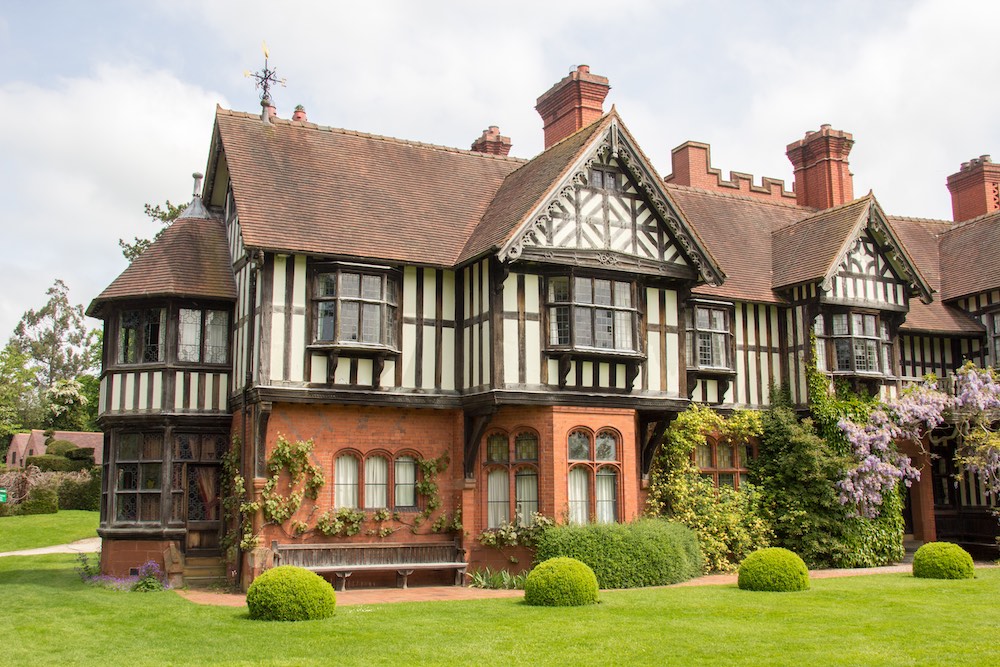 A grand Tudor-style mansion with a red brick exterior intricately detailed with white half-timbering. The residence is nestled amidst a verdant landscape, featuring a well-manicured lawn, sculpted hedges, and a vibrant wisteria vine cascading over its facade.