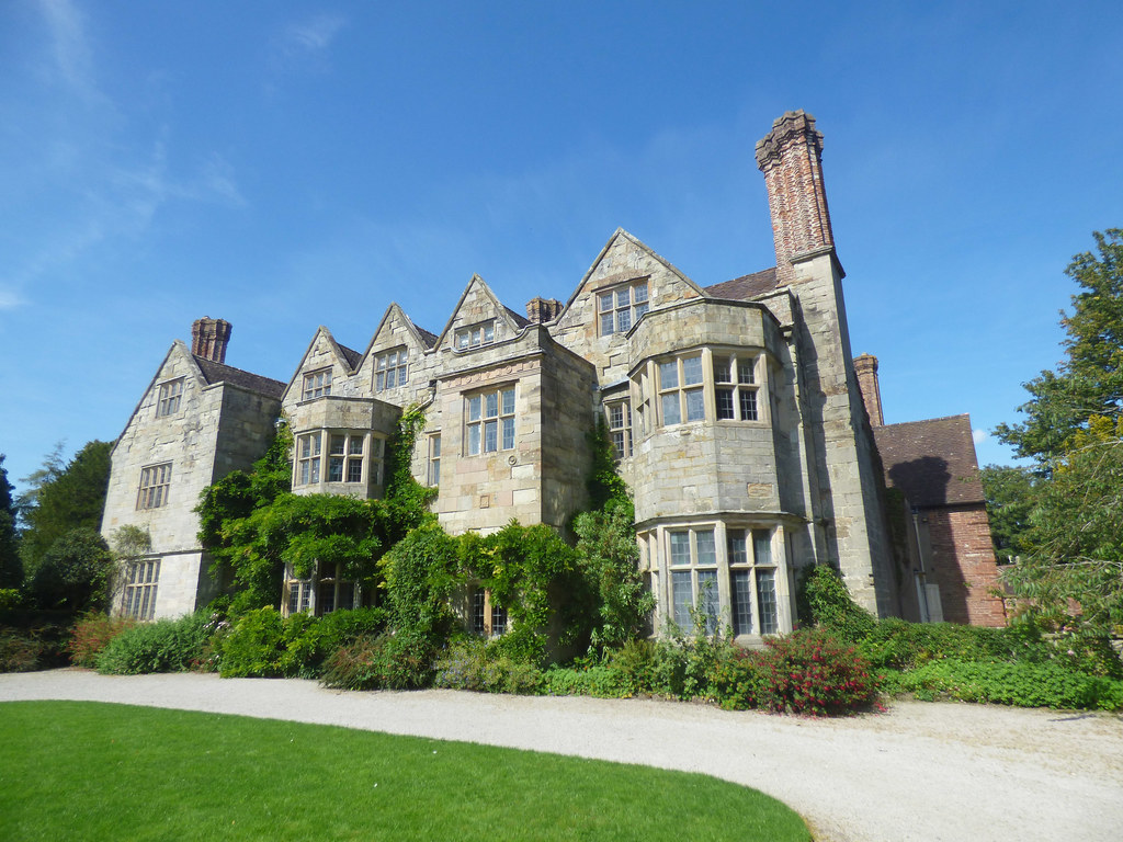 A grand historic manor house with grey stone walls, tall chimneys, and a well-manicured lawn in front, under a bright blue sky.
