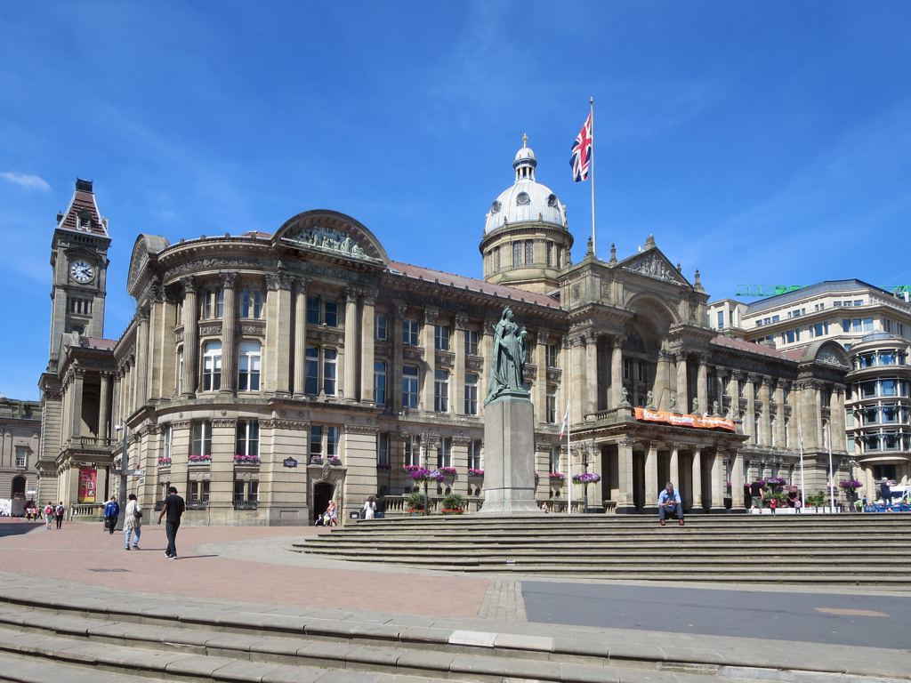 The image depicts the Birmingham Council House in Victoria Square, Birmingham, England. The grand Victorian-style building features ornate stonework and a central dome topped with a union jack flag. In front of the building, a statue of Queen Victoria stands prominently on a pedestal. The square is wet from recent rain, and several pedestrians are visible, adding a sense of daily life to the scene. The overall atmosphere is a mix of historical grandeur and typical British weather.