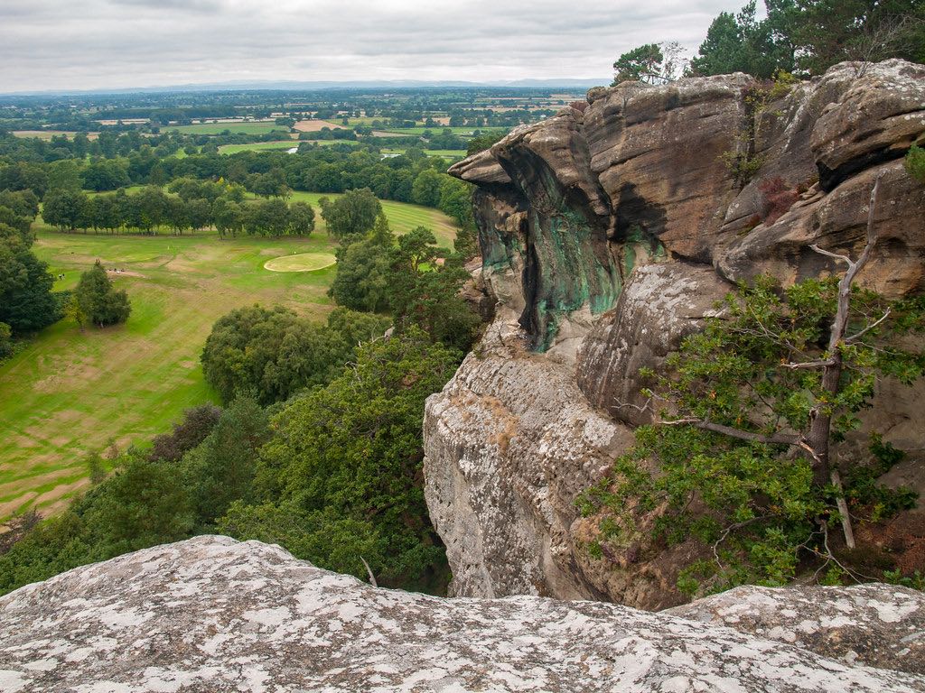 A sweeping landscape view from atop a rocky outcrop. In the foreground, weathered sandstone cliffs with patches of vegetation overlook a verdant valley below. The valley features a manicured golf course with visible greens and fairways, surrounded by dense woodland. In the distance, a patchwork of fields and forests stretches to the horizon under an overcast sky.
