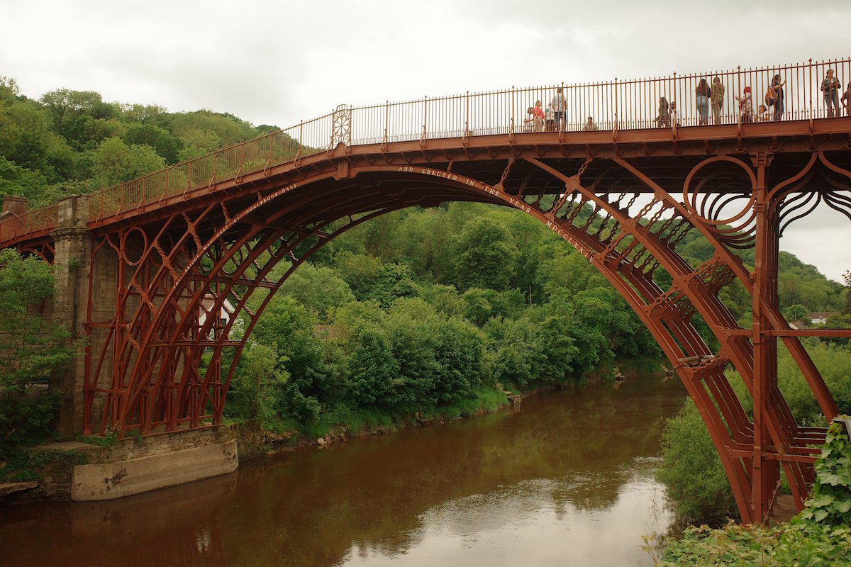A historic iron bridge spanning a wide river, with lush green trees on both sides and a few people walking across it.