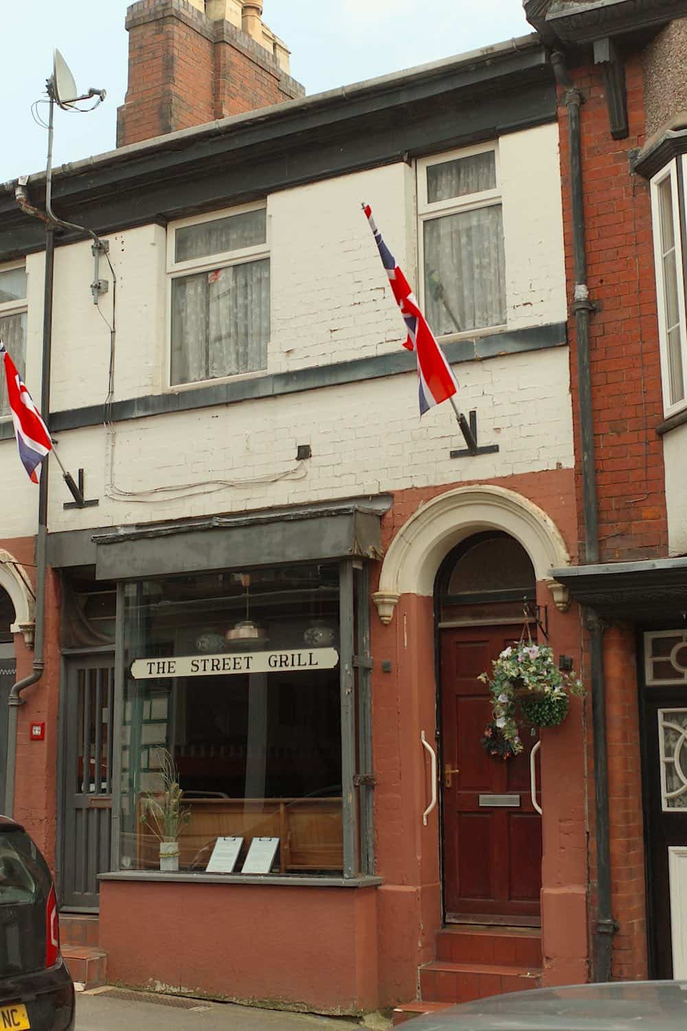 a terraced building with two union jack flags sticking out, an arched doorway with reddish brown door, a grey-framed window and door, and an old-fashioned sign on the window saying the street grill.