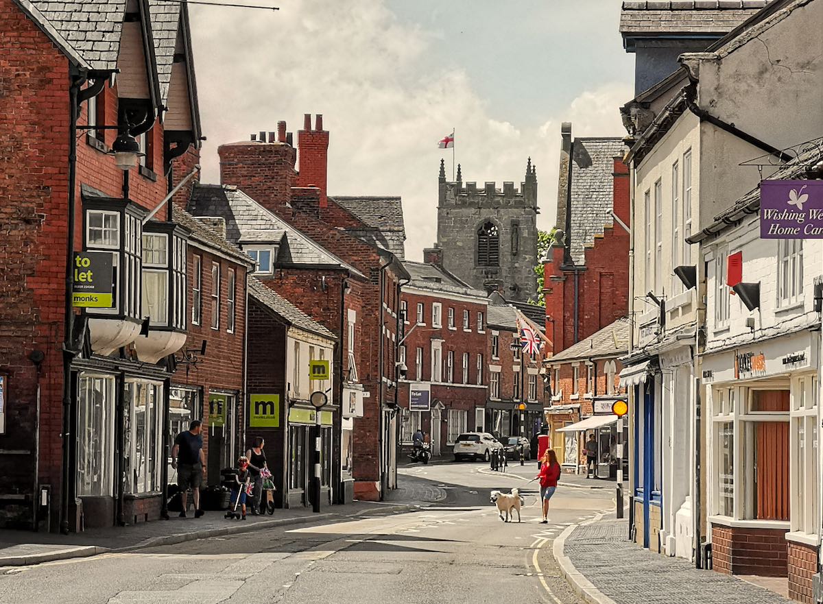 A charming English town street with red brick buildings, a church tower in the background, and Union Jack and English flags. A person in a red jacket walks a dog down the curved street, with various shops and "To Let" signs visible, and cars parked along the way.