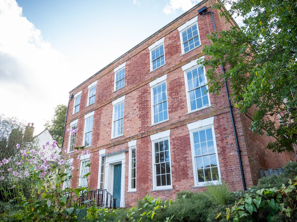 A tall, red-brick three-story house with white-framed windows and a dark teal front door. The house features traditional architectural details and is surrounded by a lush garden with purple flowers and greenery, set against a bright blue sky.