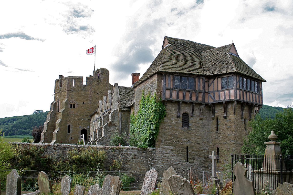 A medieval castle with a stone tower flying the English flag, connected to a half-timbered Tudor-style building. The foreground shows an old graveyard with weathered tombstones. The castle is surrounded by green hills, and the sky is cloudy. This appears to be Stokesay Castle, a fortified manor house in Shropshire, England.