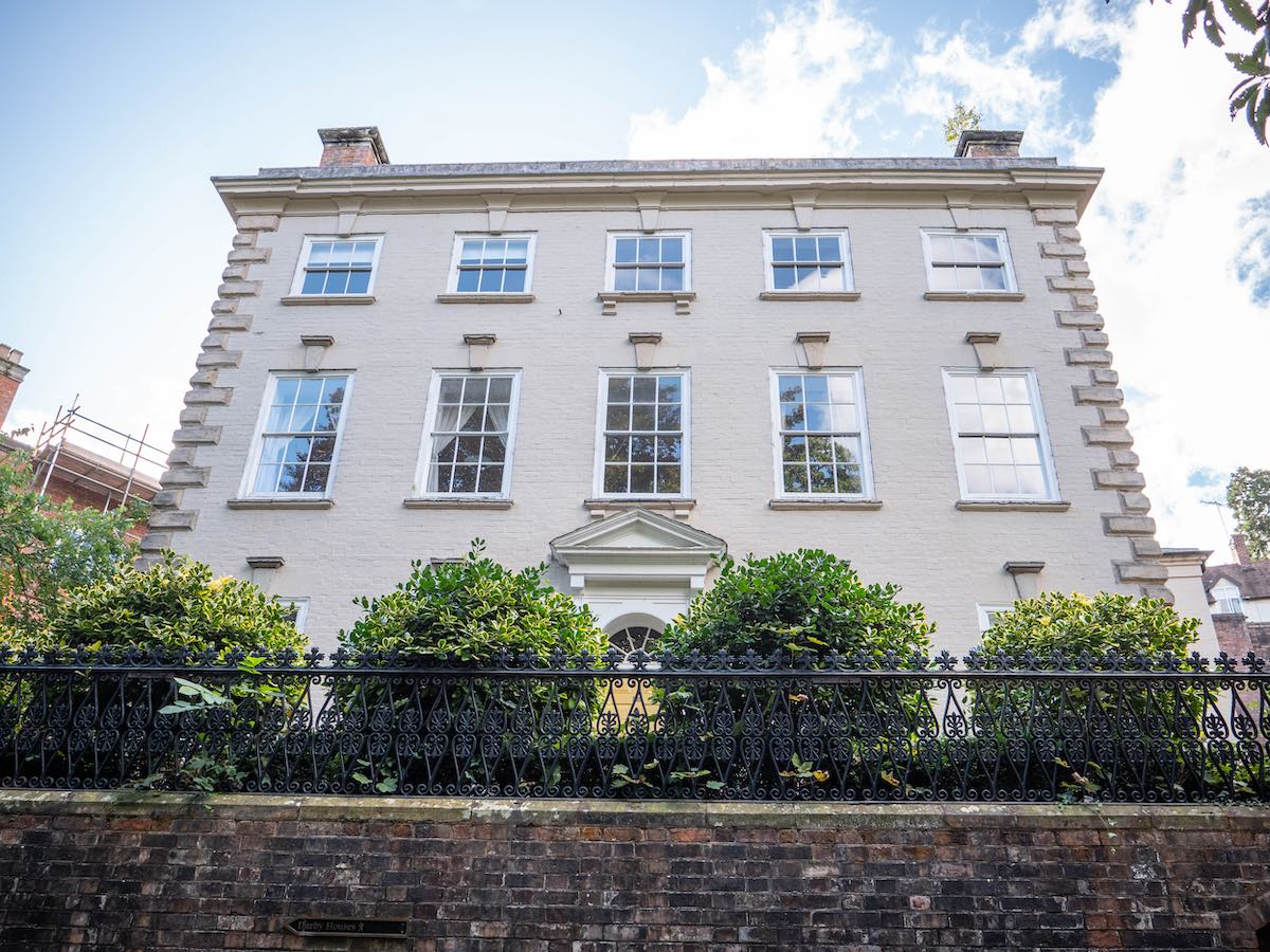 A stately Georgian-style house with white walls and large sash windows, surrounded by a black iron fence and green shrubs.