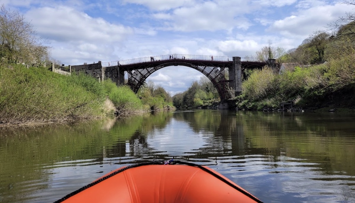 A scenic view from a boat on a calm river, looking towards a stone bridge crossing the water, with grassy banks and trees along the sides.
