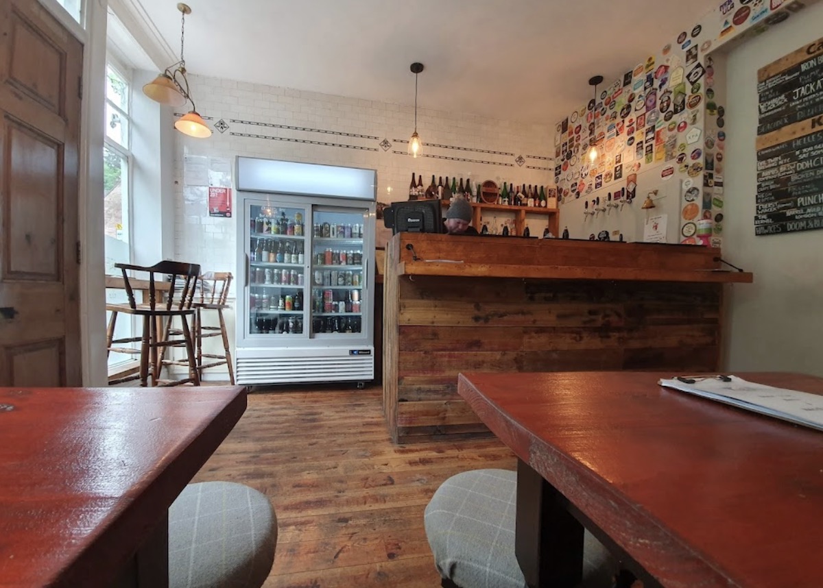 A cozy interior of a small pub with a wooden bar counter. Behind the counter, there's a man in a beanie serving customers. A large fridge filled with assorted drinks is situated to the side, and the wall behind the bar is decorated with colorful beer labels. A small table and chair set sits by a large window with natural light flooding in. Third Image (Tea Cup Ride): A colorful amusement ride featuring oversized teacups in pastel colors with heart and swirl designs. The cups spin around a large central teapot labeled "Tea Cups." Several children are seated inside the teacups enjoying the ride, surrounded by greenery and trees.