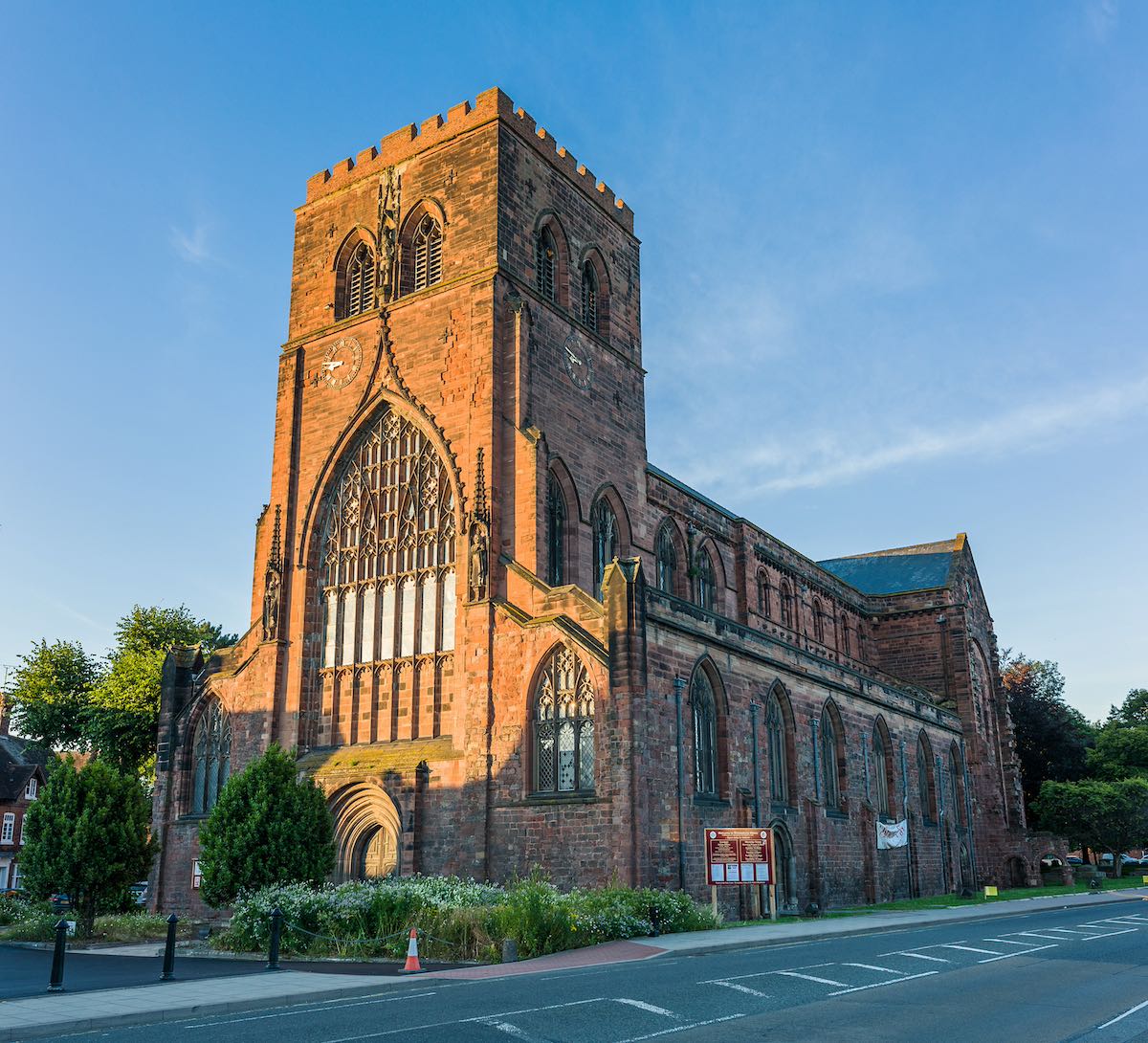 A tall, historic redbrick church with intricate Gothic architectural details, including a large stained glass window. The church is set against a clear blue sky in a peaceful landscape.