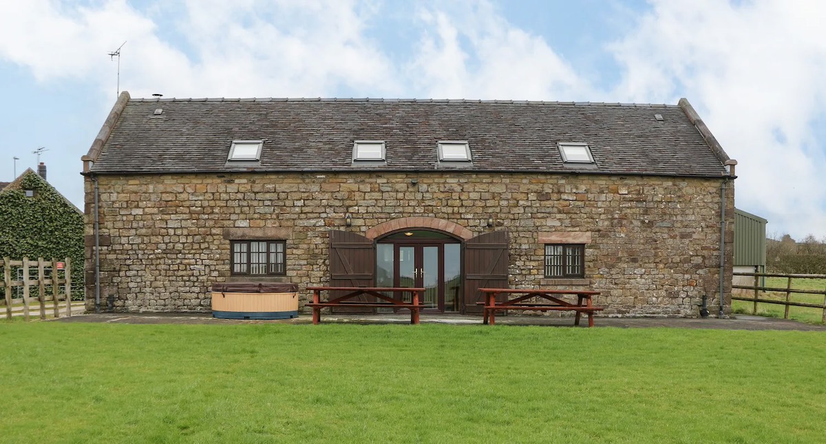 A large rustic barn with stone walls and a well-maintained grass yard. The barn features skylights and wooden doors, and picnic benches are placed in front of the structure.