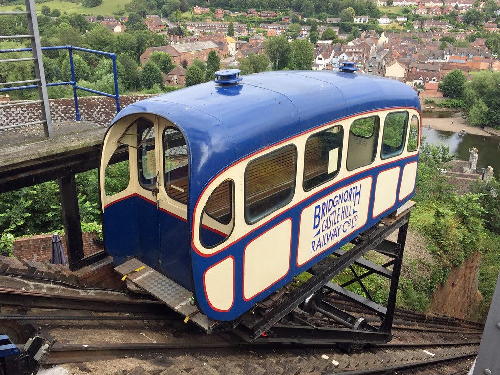 A blue cable car or funicular labeled "Bridgnorth Castle Hill Railway Co Ltd" sits on an inclined track overlooking a town with red-roofed buildings and green countryside in the background.