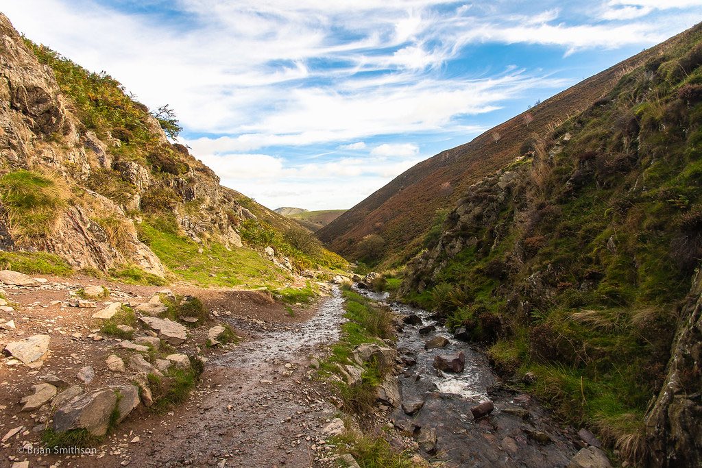 A scenic rocky valley with a small stream running through it. The valley is flanked by steep hillsides covered in vegetation. A narrow path runs alongside the stream, and wispy clouds can be seen in the blue sky above.