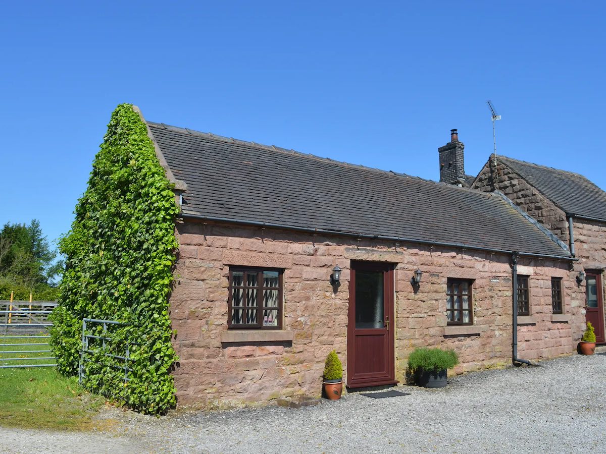 A traditional stone-built cottage with red doors and windows. The cottage has ivy growing on its walls, with a clear blue sky in the background.