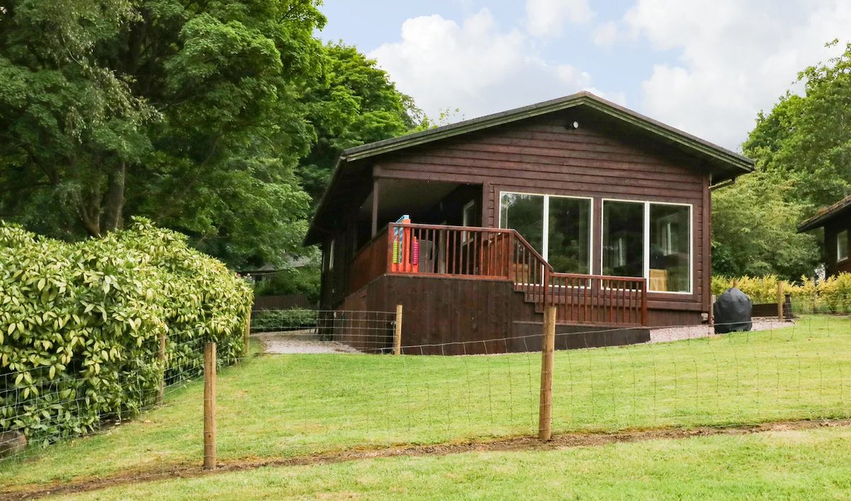 A cozy wooden cabin located in a peaceful, green forest setting. The cabin has a deck with outdoor seating and is enclosed by a simple wooden fence.