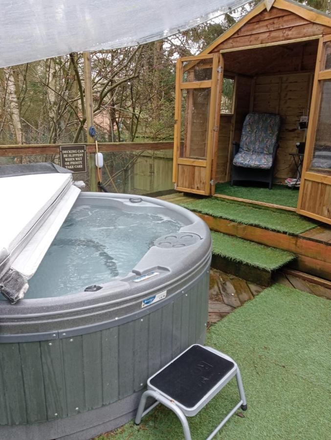 A cozy wooden structure featuring a private hot tub on a green artificial turf, with a small garden shed in the background. The tub is placed on a slightly raised platform with steps.