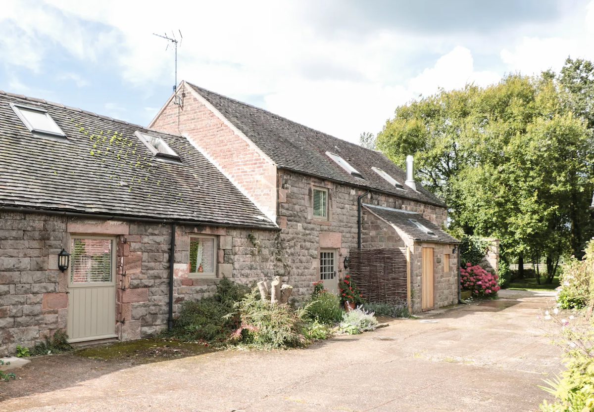 A charming stone-built cottage with sloped roofs, set amidst trees and an open yard. The cottage has several windows and a simple wooden door.