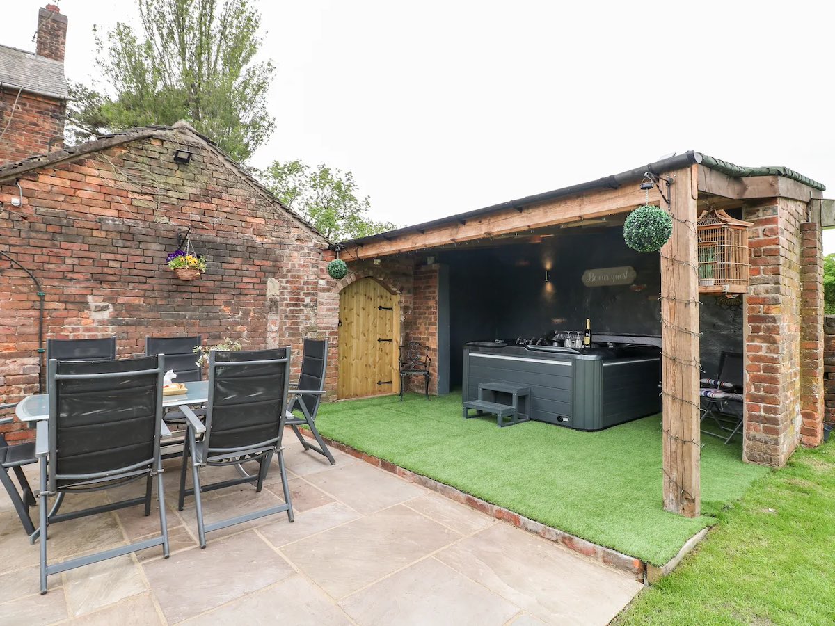 A brick-walled house with an outdoor patio featuring a large hot tub and a dining area. The patio is covered by an open wooden shelter, with artificial grass on the floor.